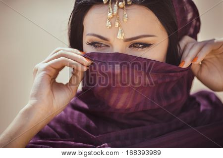 Beautiful Arabian woman portrait outdoors. Young Hindu woman. Portrait of beauty Indian model with bright make-up who hiding her face behind the veil standing over gold desert background.