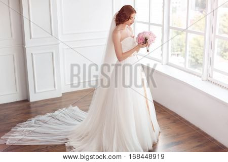 Bride in white wedding dress and long veil with bouquet of flowers indoors. Young gorgeous fashion model posing in white interior studio's room near big window in full lenght