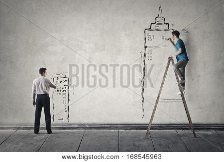 Men drawing buildings on grey wall, with the help of a ledder 