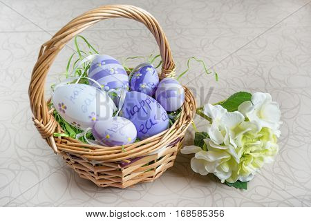 Easter wicker basket with colored eggs and a white flower on white board.