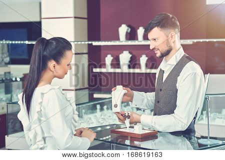 Jeweler young brunet man with beard showing for female buyer necklace of gold jewelry set with black stones. Side view of woman in blue blouse choosing present and want buying jewelry for herself.