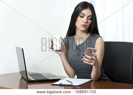 Young business woman at her office. She is holding a cup of coffee in one hand and a smart phone in the other hand.