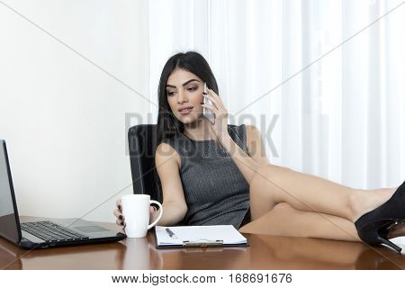 Business woman relaxing with her legs on the table. she is holding a cup of hot drink while she is talking on her mobile phone.