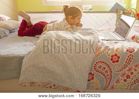 Little girl lying in bed and watching cartoons with a laptop in his bedroom. Sun light entering by window