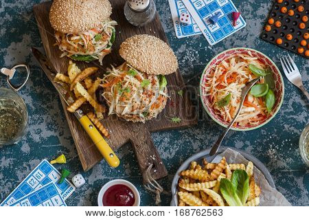 Hamburgers with grilled chicken and cole slaw on a wooden board on the table with cards and bingo chips top view. Flat lay