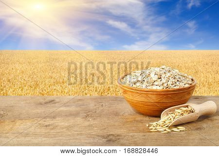 Oat flakes in bowl. Grains of oat in scoop and oatmeal in bowl on wooden table with field on the background. Ripe field, blue sky with beautiful clouds and sun. Uncooked porridge