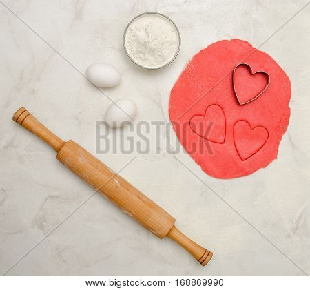 Red dough with a cut out heart eggs flour and rolling pinon a white table. Top view space for text