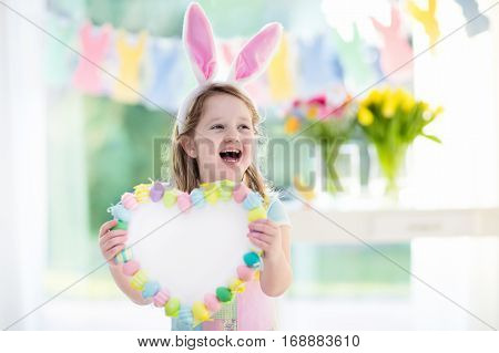 Happy little girl in bunny ears holding a heart board with colorful Easter eggs. Kids celebrate Easter. Children having fun on Easter egg hunt. Pastel bunny banner and flowers. Space for your text.