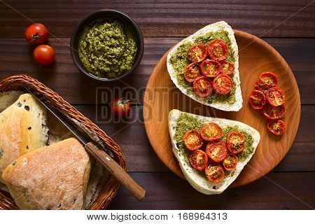 Olive bread roll halves spread with basil pesto and topped with roasted cherry tomato halves served on wooden plate photographed overhead on dark wood with natural light
