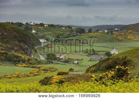 North Coast Scotland - June 6 2012: Hilly green country side in the Farr region of Northern Scotland. White houses sprinkled among green meadows brown rocky hills dark green trees and yellow broom flowers. Road meanders through the scene under dark skies.