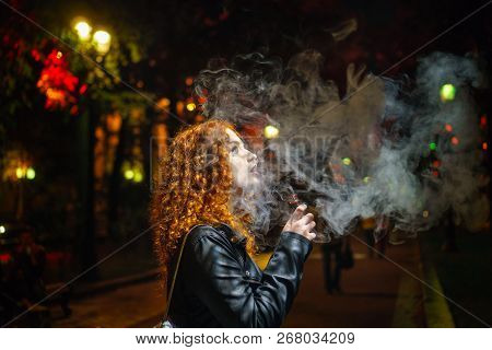 Young Red-haired Girl Thoughtfully Smokes An Electronic Cigarette. She Walks Alone On Streets Of Nig