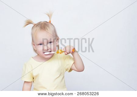 Little Blue-eyed Girl Blonde With Pigtails On Her Head Brushing Teeth With A Toothbrush And Looking 