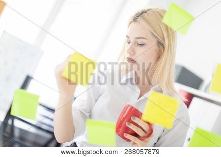 A Young Girl Stands Near A Transparent Board With Stickers. Shes Holding A Cup And A Marker.