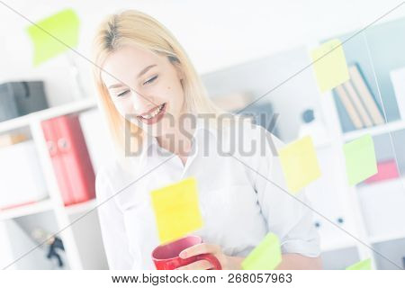 A Young Girl Stands Near A Transparent Board With Stickers. Shes Holding A Cup And A Marker.