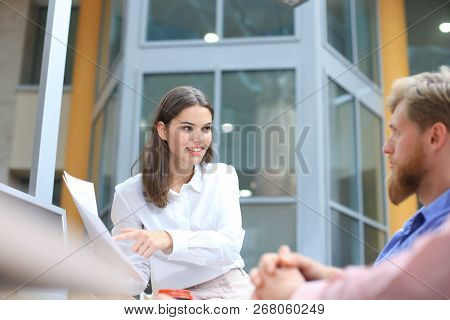 Group Of Young People In Casual Wear Sitting At The Office Desk And Discussing Something While Looki