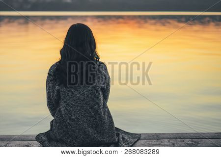 Back View Of Long Hair Brunette Girl Sitting On The Pier Near Lake At Sunset Time.