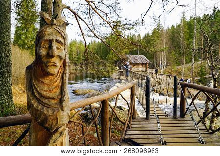 Carved Wooden Sculpture Of An Old Woman Near The Pedestrian Suspension Bridge Over A Waterfall On Th