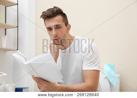 Young Man With Book Sitting On Toilet Bowl In Bathroom