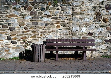 A Plastic Park Bench And A Garbage Bin Stand In Front Of A Striking And Rugged Wall Of Fieldstone.