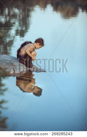 Caveman Boy Sitting On The Rock And Looking At Him Self In The Water Reflection In Lake. Evolution S