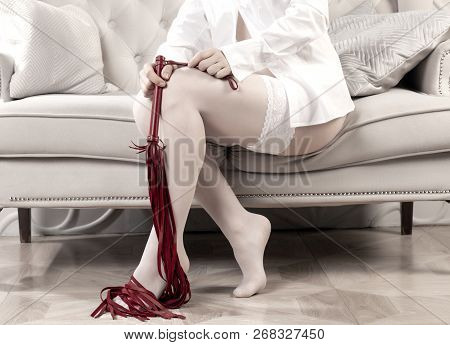 Woman In White Stockings Holds A Whip Sitting On The Couch. Female Legs And Whip