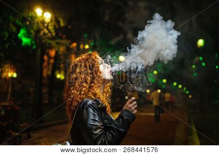 Young Red-haired Girl Dreamily Smokes An Electronic Cigarette. She Walks Alone On Streets Of Night C