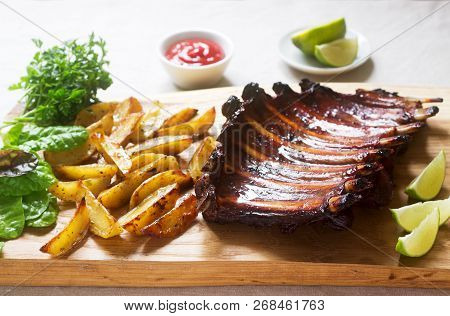 Homemade Baked Meat Ribs Served With French Fries, Herbs, Lime And Ketchup On A Wooden Board. Rustic