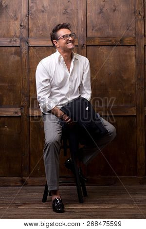 Optimistic Relaxed Middle Aged Man Sitting On The Bar Stool With A Jacket In His Hand And Putting Hi