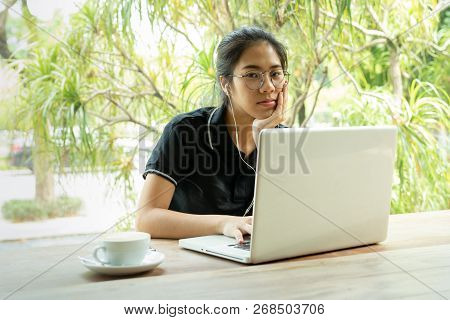 Asian Woman Student Working On Laptop With Earphone Looking At Camera.
