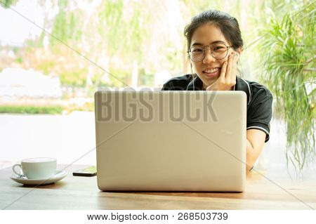 Asian Woman Student Working On Laptop With Earphone Looking At Camera.