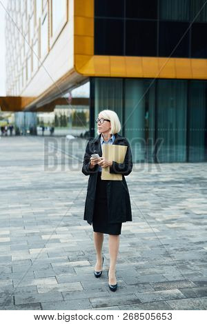 Mature elegant businesswoman in black coat moving down trottoire in urban environment