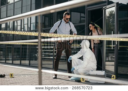 Detectives And Criminologist  In Protective Suit And Latex Gloves Communicating At Crime Scene