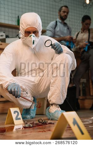 Concentarted Forensic Investigator Examining Evidence With Magnifying Glass At Crime Scene With Coll