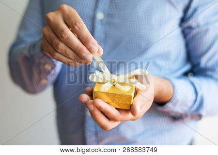 Close-up Of Man Untying Bow On Small Yellow Box. Unrecognizable Man Opening Christmas Gift. Birthday