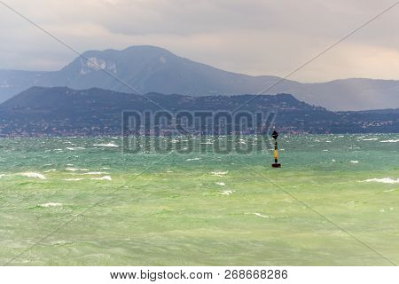 Black And Yellow Buoy In Turbulent Blue Green Waves On Lago Di Garda Lake, Windy, Cloudy, Foggy Weat