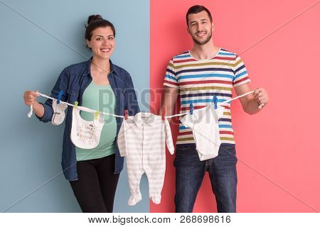 Beautiful pregnant woman and her husband expecting baby holding baby bodysuits and smiling over colorful background