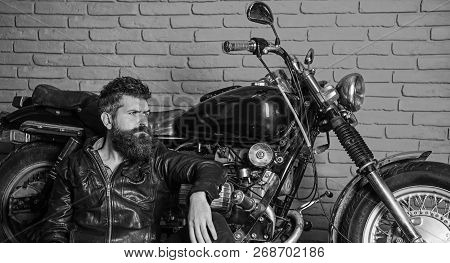 Man With Beard, Biker In Leather Jacket Near Motor Bike In Garage, Brick Wall Background. Bikers Lif