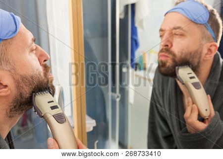 Focused Bearded Man Shaving His Beard Using Electric Razor. Taking Care Of Facial Hair, Wearing Slee