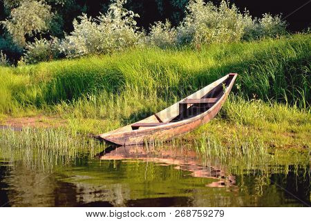 Old Wooden Boat On The River Bank Close Up Against The Background Of Green Grass And Bushes, Reflect