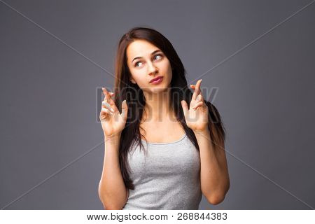 Body language and emotions concept. Glad dark haired beautiful satisfied girl beggs God to help her, keeps fingers crossed, wishes good luck, prays indoor isolated over gray studio background.