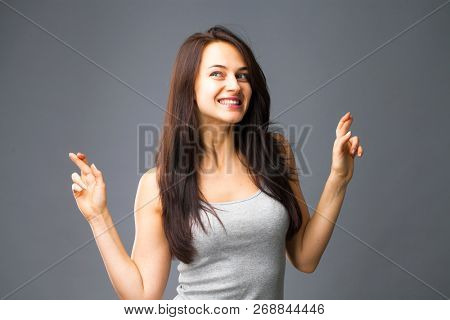 Body language and emotions concept. Glad dark haired beautiful satisfied girl beggs God to help her, keeps fingers crossed, wishes good luck, prays indoor isolated over gray studio background.