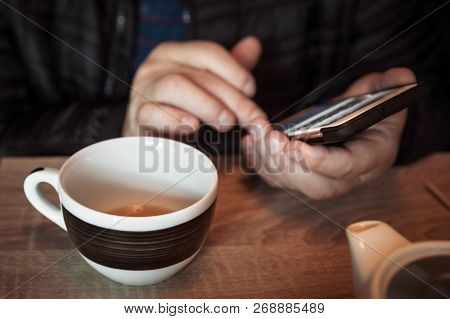 A Man Is Resting In A Cafe, Drinking Tea, Reading And Writing Messages On A Smartphone. Work In The 