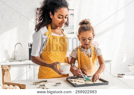 African American Mother And Daughter In Yellow Aprons Applying Oil On Unbaked Cookies In Kitchen