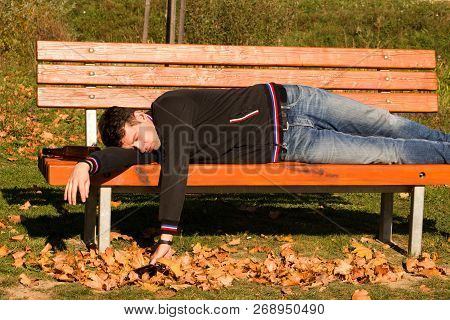 A Drunk Young Man Sleeping On A Bench In The Day.a Young Man Holding A Bottle Of Alcohol In Her Hand