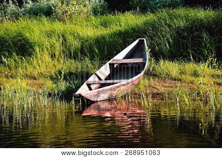 Old Wooden Boat On The River Bank Close Up Against The Background Of Green Grass And Bushes, Reflect