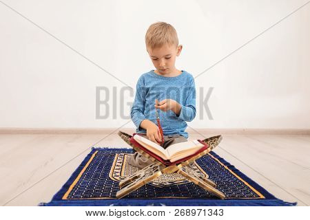 Little Muslim Boy With Misbaha And Koran Praying On Rug Indoors