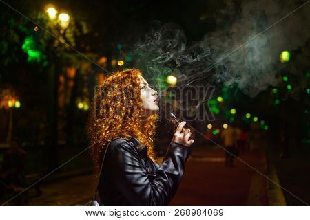 Young Red-haired Girl Smokes An E-cigarette. She Walks Alone On Streets Of Night City. Non-nicotine 