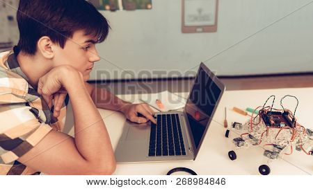 Upset Teenager In Shirt Programming Robot At Home. Young Man In Shirt. Using Laptop. Modern Hobby Co