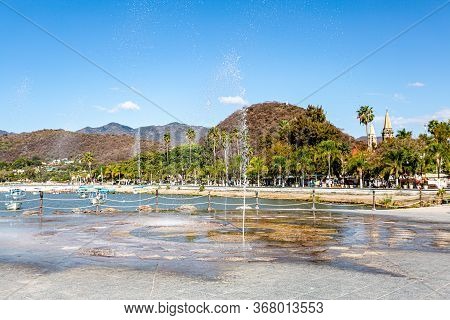 Fountain Spraying The Ground With Jets Of Water On The Promenade Of Lake Chapala With The Town In Th