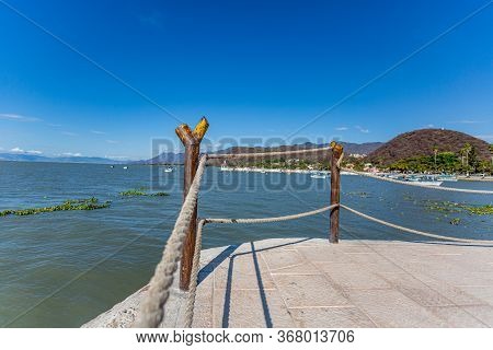 Boardwalk With A Fence With Wooden Posts And Ropes Next To Lake Chapala With Calm Waters, The Pier A